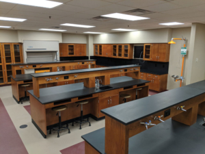 biology lab room shot. The room has three large, 8-person desks with water and gas access. A yellow emergency shower stands against the cabinet-lined wall.